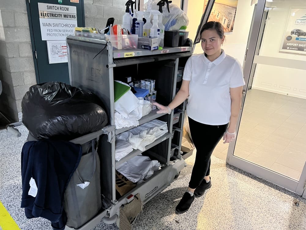 Cleaning staff member at Honeysuckle Executive Apartments standing beside fully stocked housekeeping trolley cart